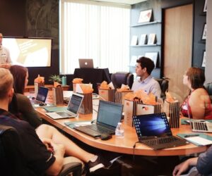 Schulungen für Mitarbeitende sind eine langfristige Investition. Um wirkungsvoll zu sein, sie gut geplant und die Lernergebniosse nachgehalten werden. Menschen sitzen mit Laptops um einen Konferenztisch und bekommen von einem Trainer eine Schulung.