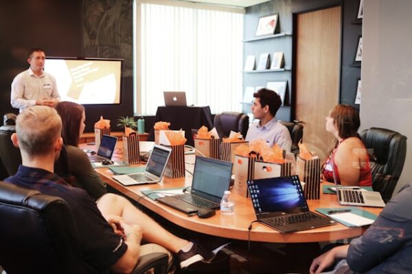 Schulungen für Mitarbeitende sind eine langfristige Investition. Um wirkungsvoll zu sein, sie gut geplant und die Lernergebniosse nachgehalten werden. Menschen sitzen mit Laptops um einen Konferenztisch und bekommen von einem Trainer eine Schulung.