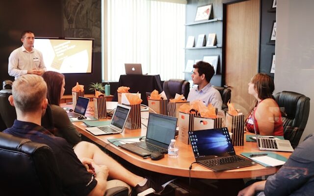 Schulungen für Mitarbeitende sind eine langfristige Investition. Um wirkungsvoll zu sein, sie gut geplant und die Lernergebniosse nachgehalten werden. Menschen sitzen mit Laptops um einen Konferenztisch und bekommen von einem Trainer eine Schulung.
